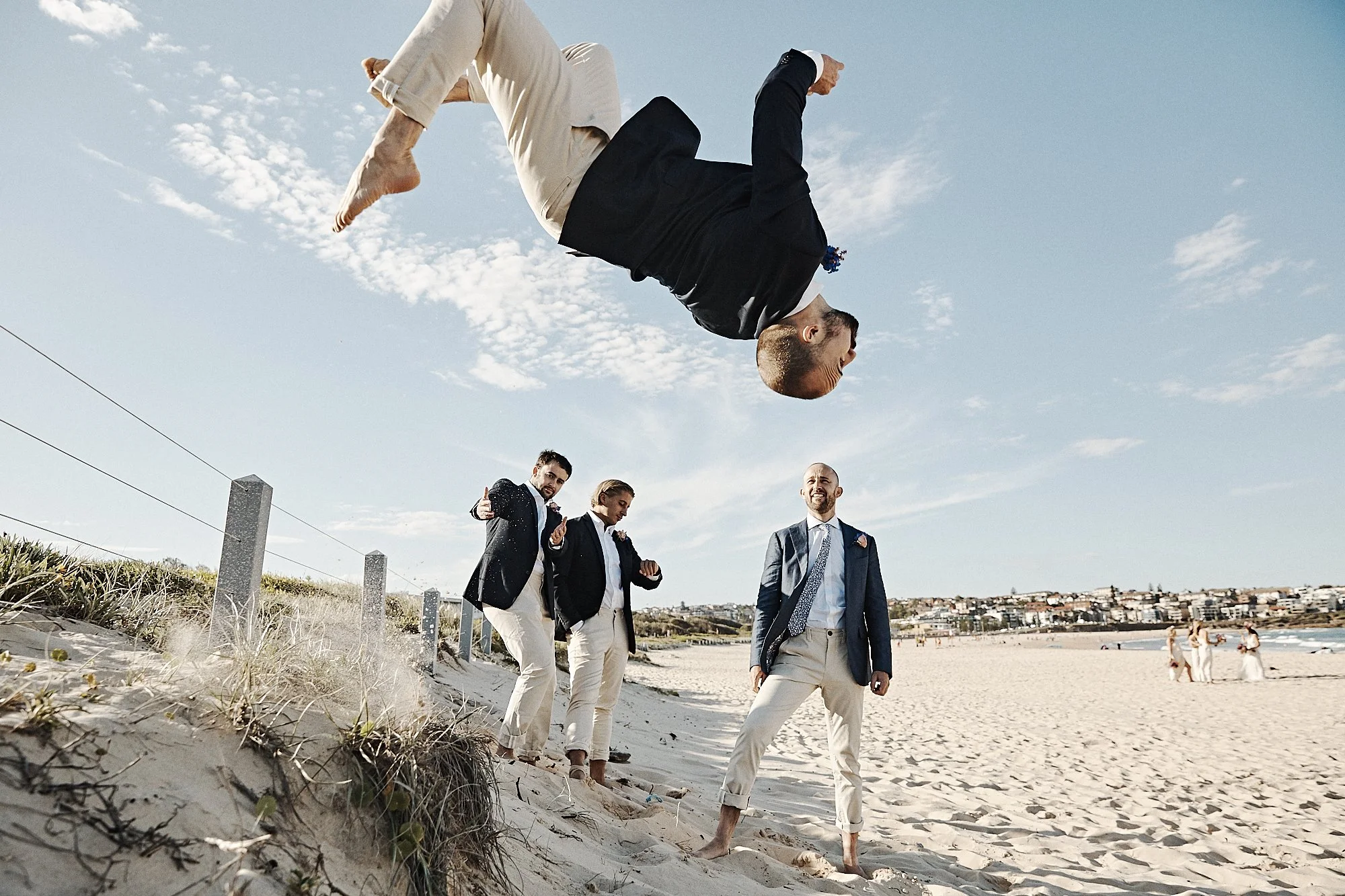 A group of five men dressed in formal suits walking barefoot on a sandy beach with a cityscape in the background. One man is mid-air, jumping or flipping, and others are walking with confident poses. The sky is mostly clear with some clouds.