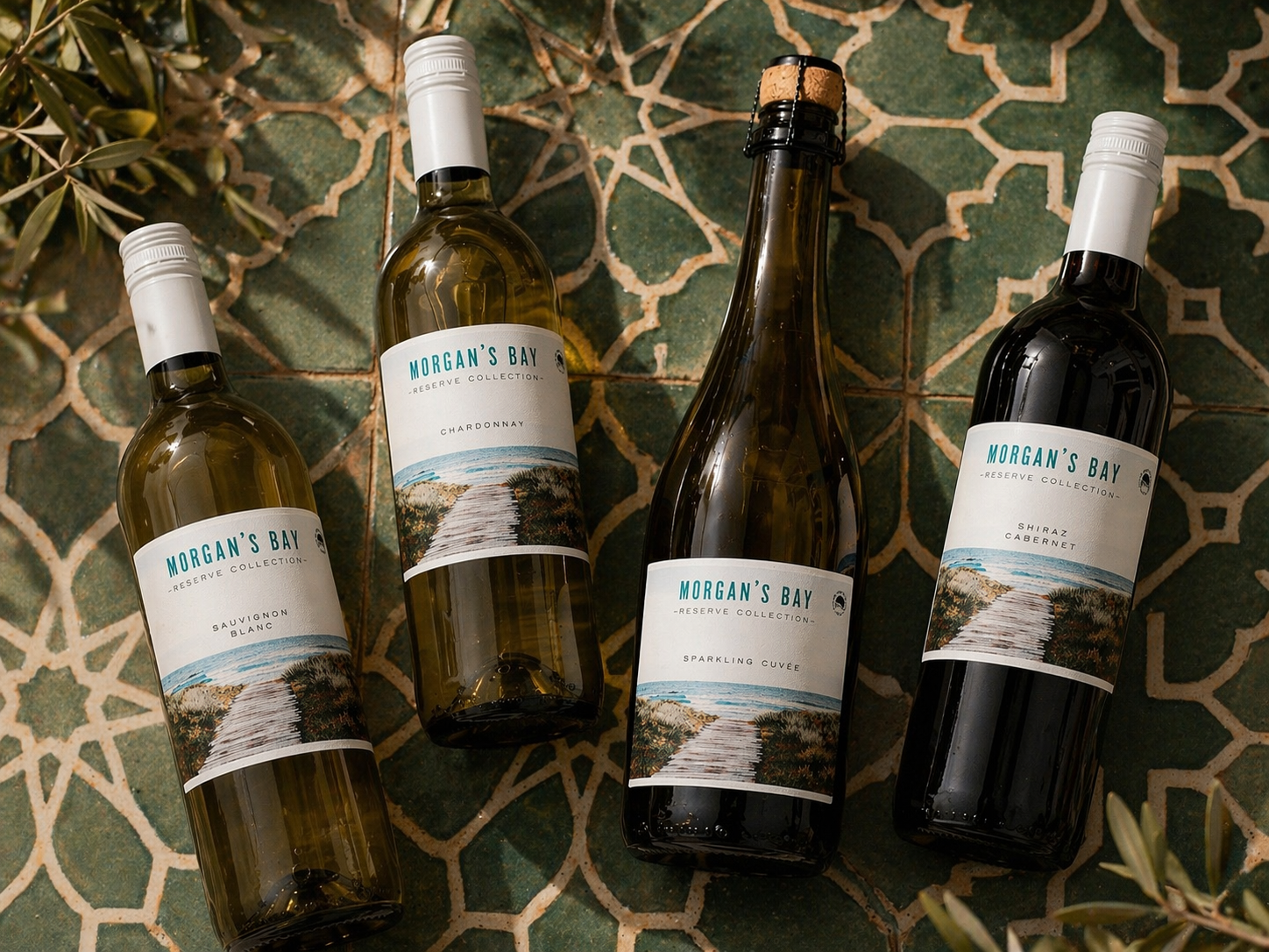 Four bottles of Morgan's Bay Reserve Collection wine placed on a patterned tile floor, with two white wine bottles, one sparkling wine bottle, and one red wine bottle, each with a scenic beach-themed label.