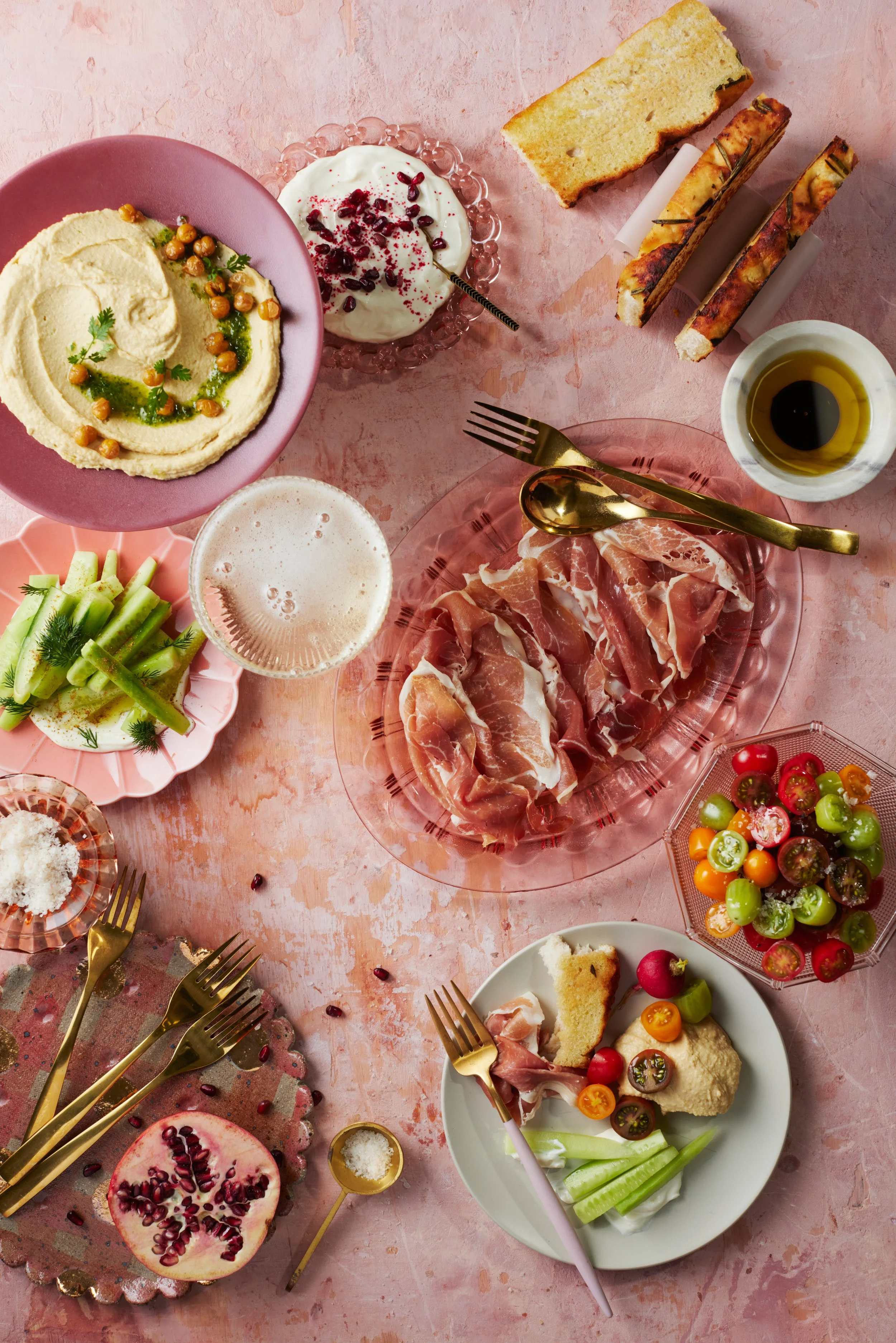 A colorful spread of food on a pink table includes hummus, a yogurt dish topped with pomegranate seeds, sliced breadsticks, olive oil, prosciutto, fresh vegetables, radish slices, and a fruit platter with pomegranate