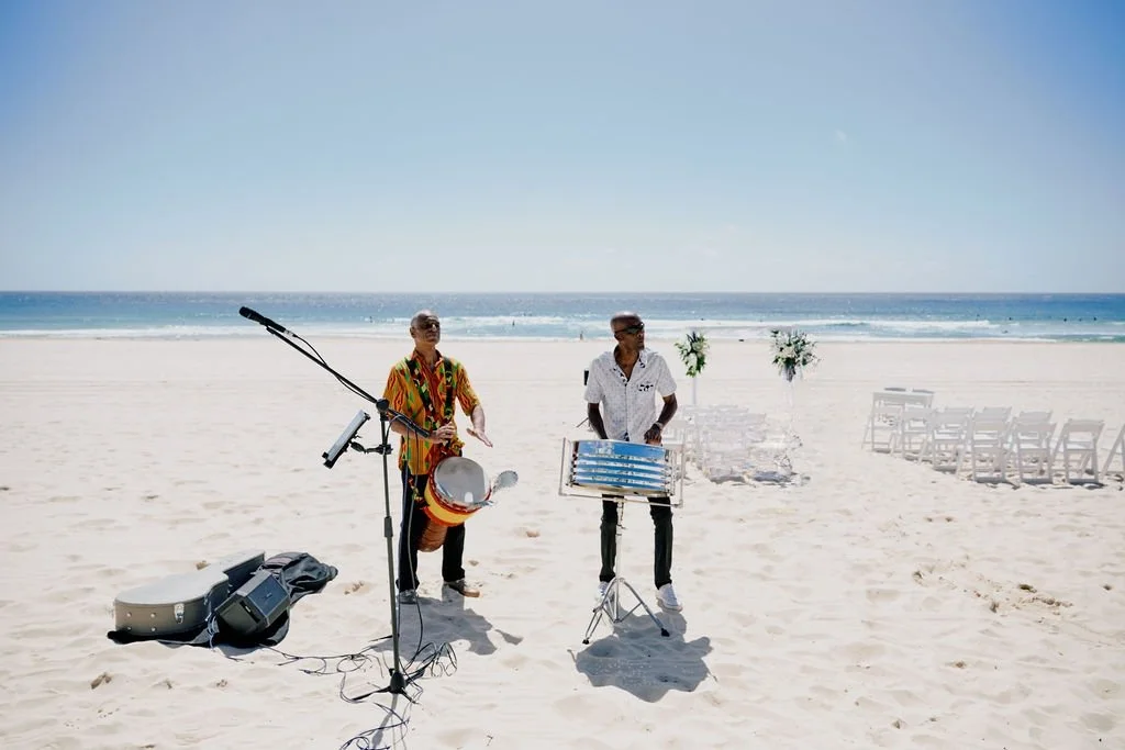 Two musicians playing instruments on a beach with chairs and floral arrangements in the background.