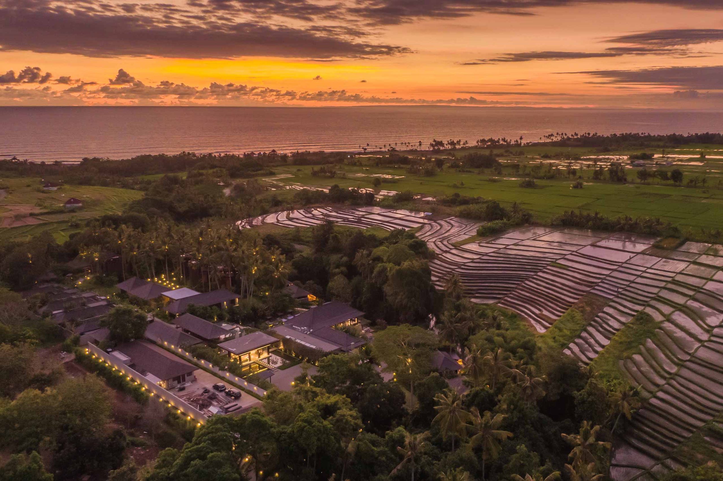 Sunset over rice terraces and a coastline with a few buildings and trees, illuminated with outdoor lighting.