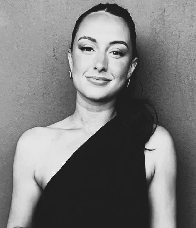 Black and white portrait of a woman with dark hair styled back, wearing earrings, and a one-shoulder black dress, standing against a plain wall.