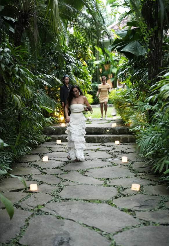A woman in a white strapless dress walking down a stone pathway decorated with small candles, surrounded by lush green tropical plants, with two other women in the background.