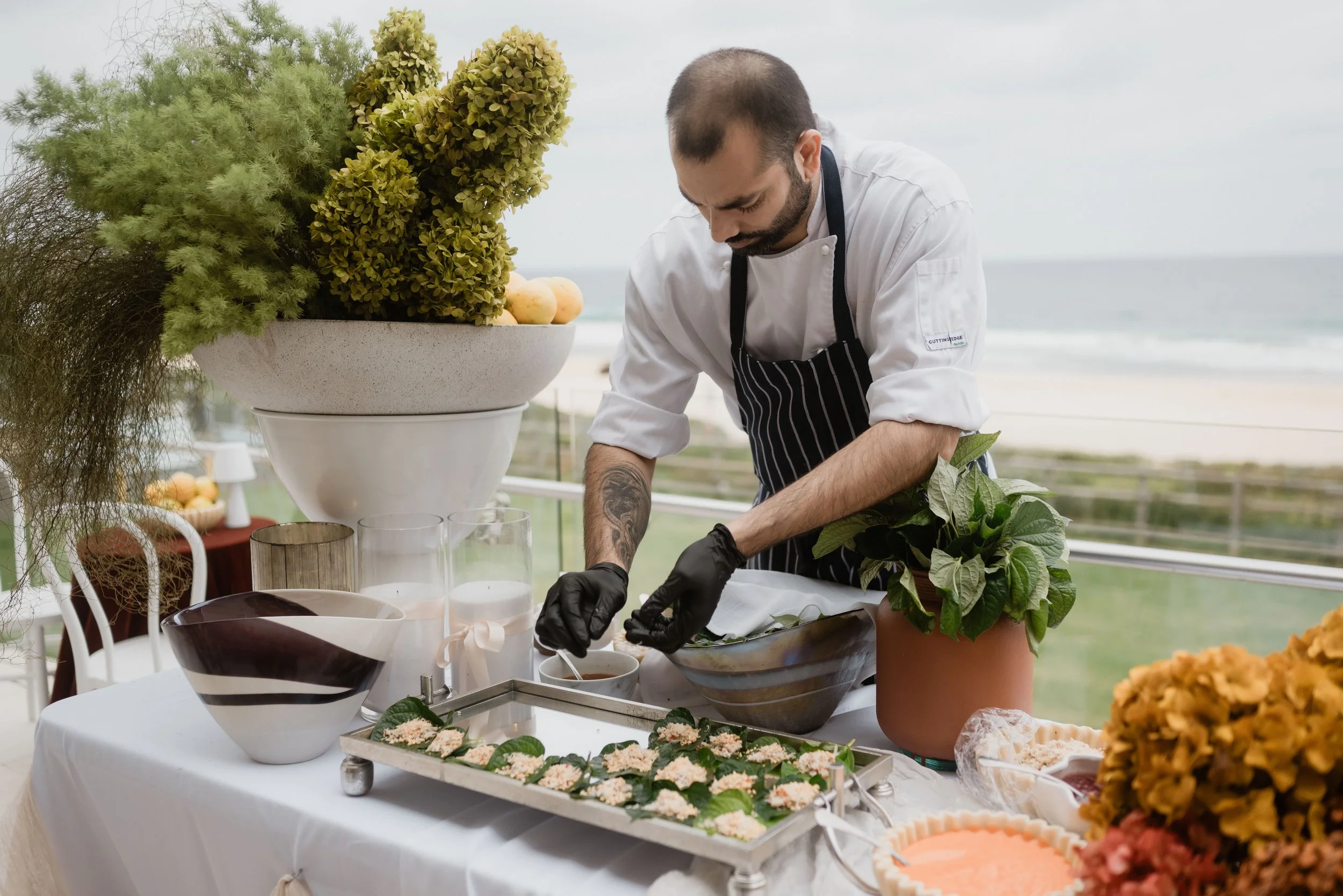 A chef wearing black gloves and a striped apron preparing food on an outdoor table filled with various dishes and ingredients, with a large bowl of decorative plants and the ocean in the background.