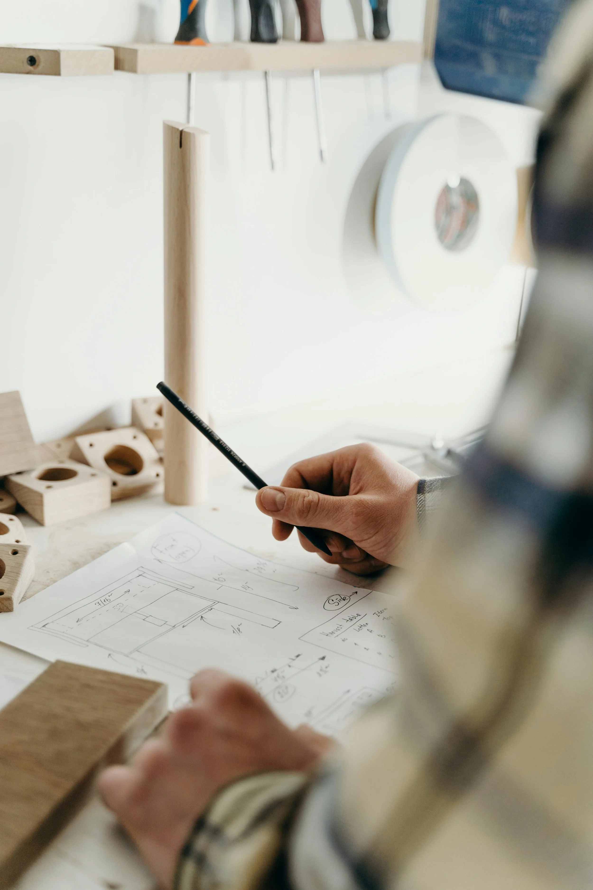 A person working on a woodworking project, holding a pen over a sketch with measurements and drawings, on a table with wooden pieces and a handsaw.