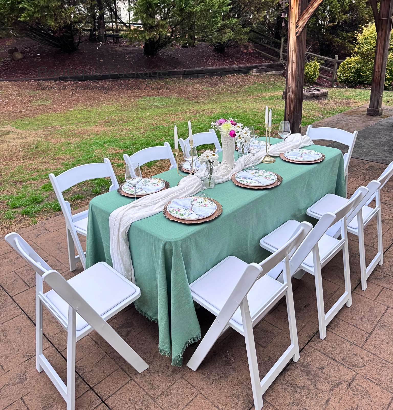 Outdoor dining table set with a green tablecloth, white chairs, floral plates, wine glasses, and floral centerpieces on a patio surrounded by grass and trees.