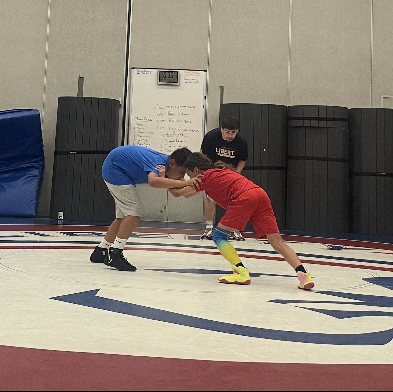 Two young boys are wrestling on a wrestling mat, with a coach observing behind them.