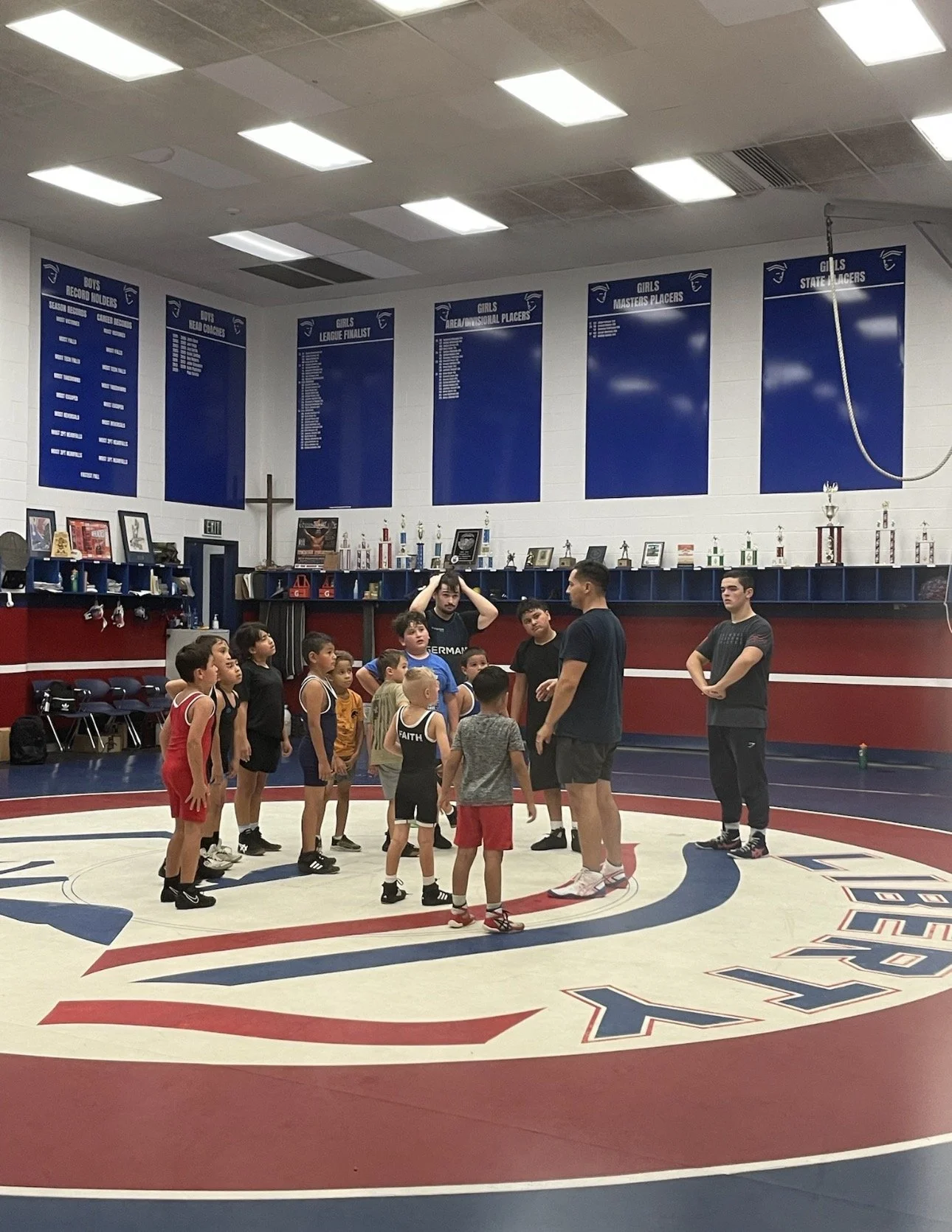 A group of young children listening to a coach during a wrestling practice or class in a wrestling gym, with trophies and posters in the background.