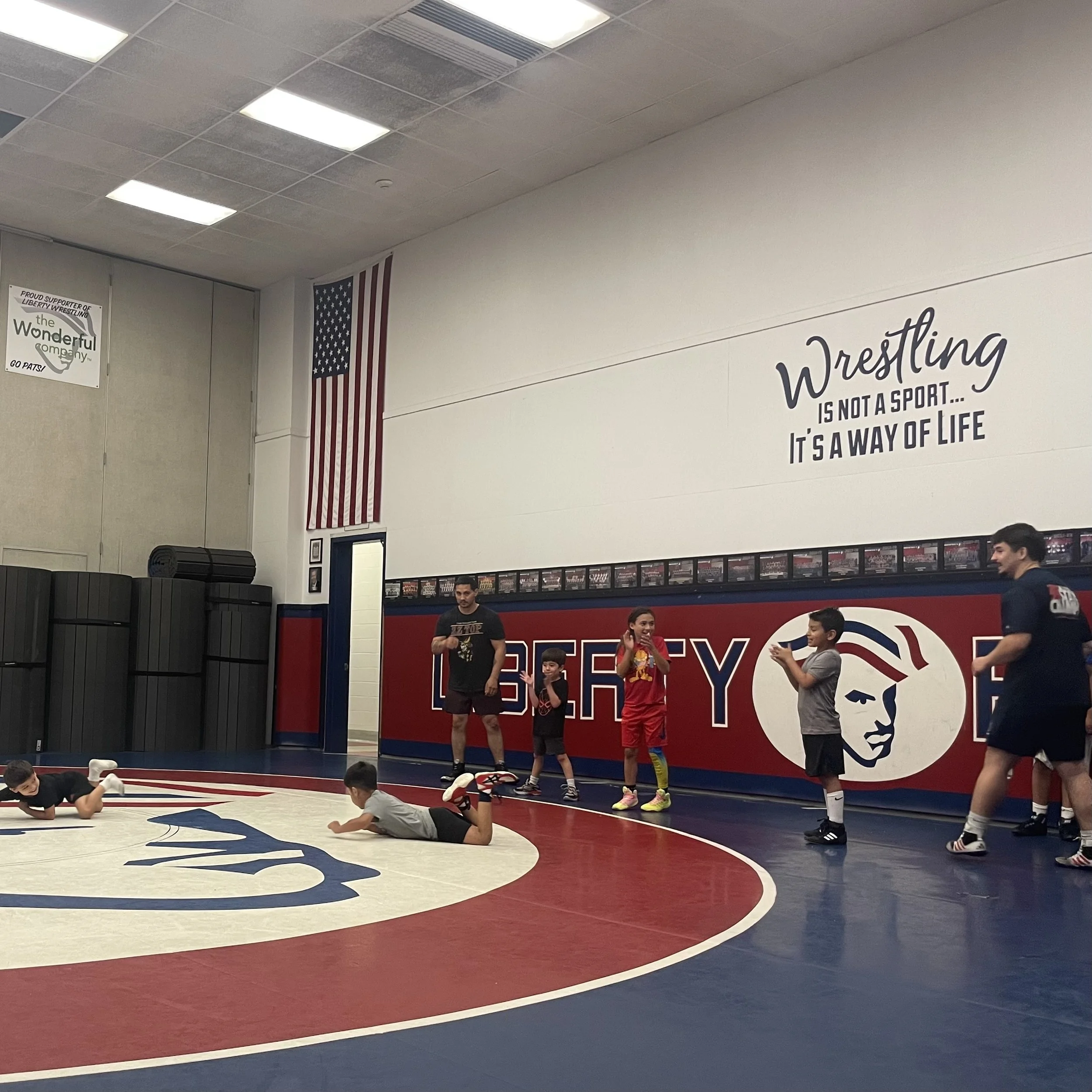 Children and adults in a wrestling gym, some stretching and warming up on a red, blue, and white mat, with a large American flag and wrestling posters on the walls.