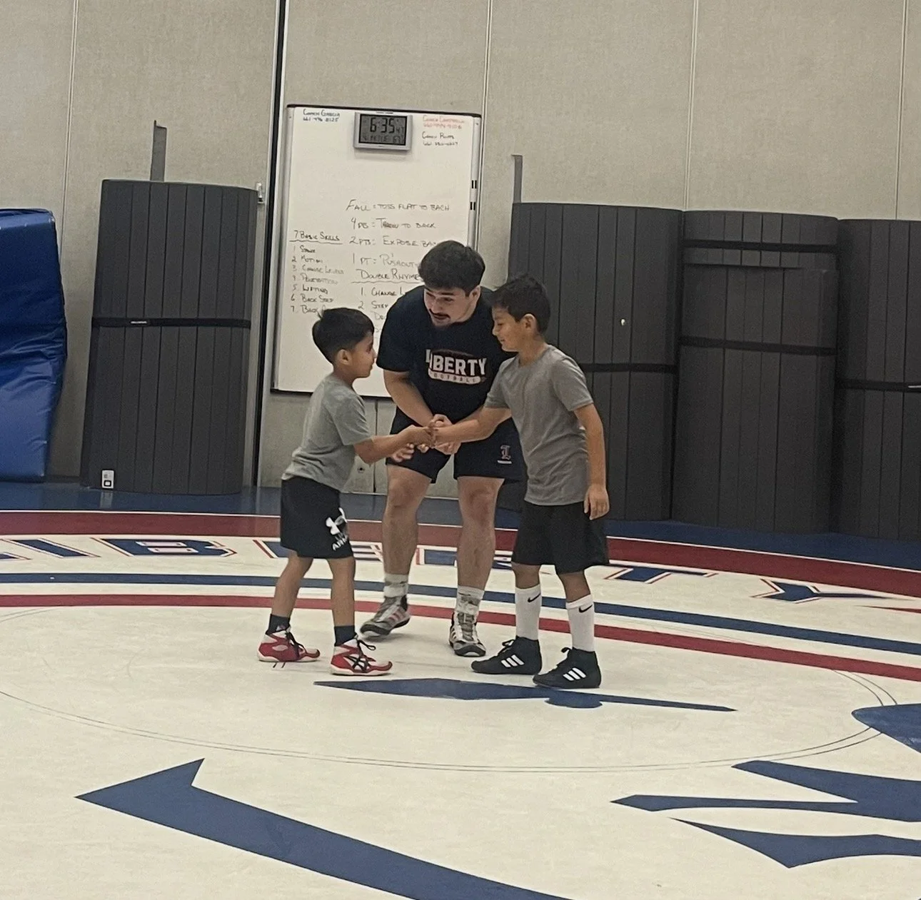 A coach training two young boys on a wrestling mat.