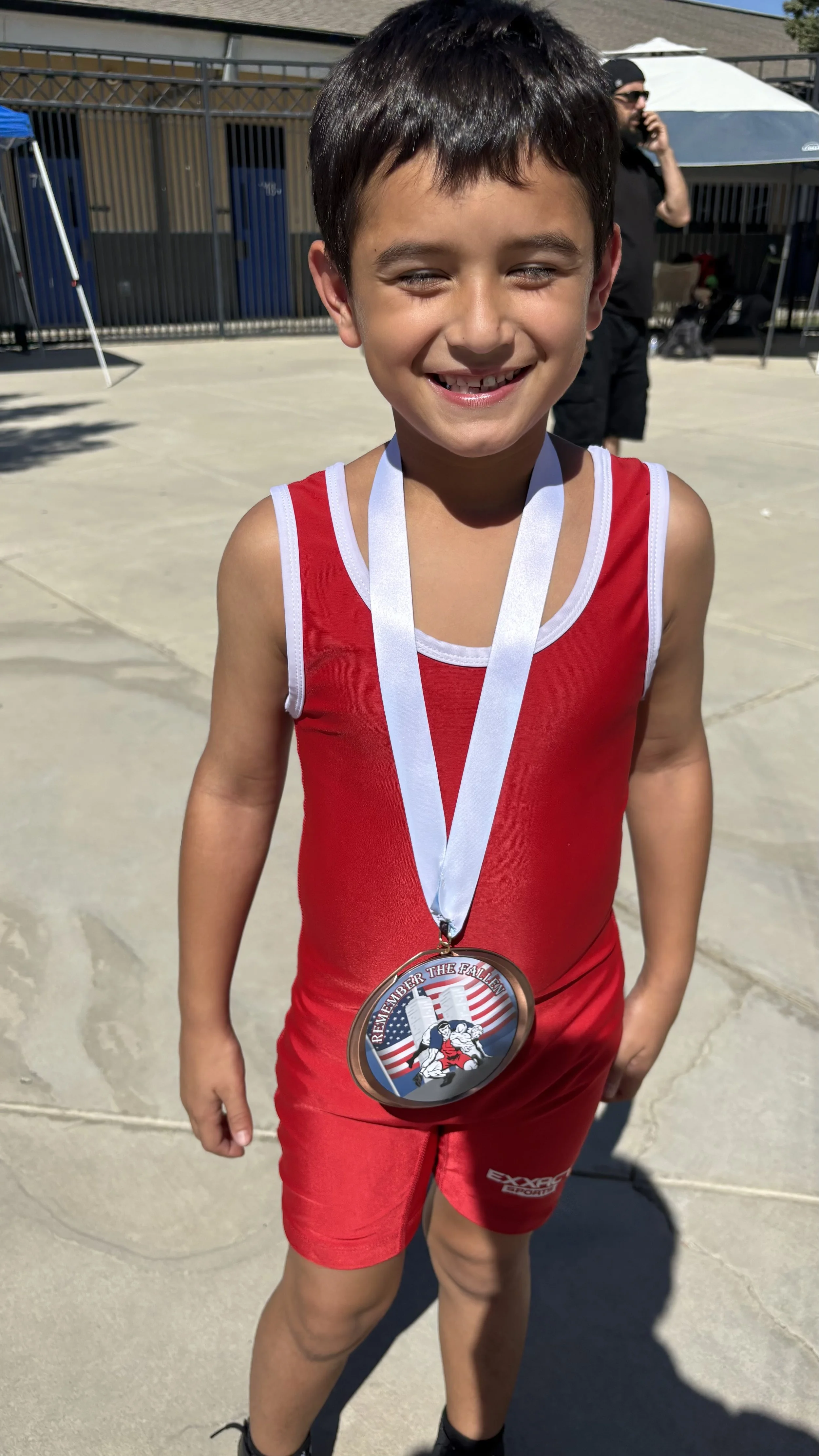 A young boy in a red athletic uniform with white trim, smiling and showing a missing tooth, is wearing a medal around his neck. The background shows an outdoor area with a fence, a person talking on a cell phone, tents, and other people.