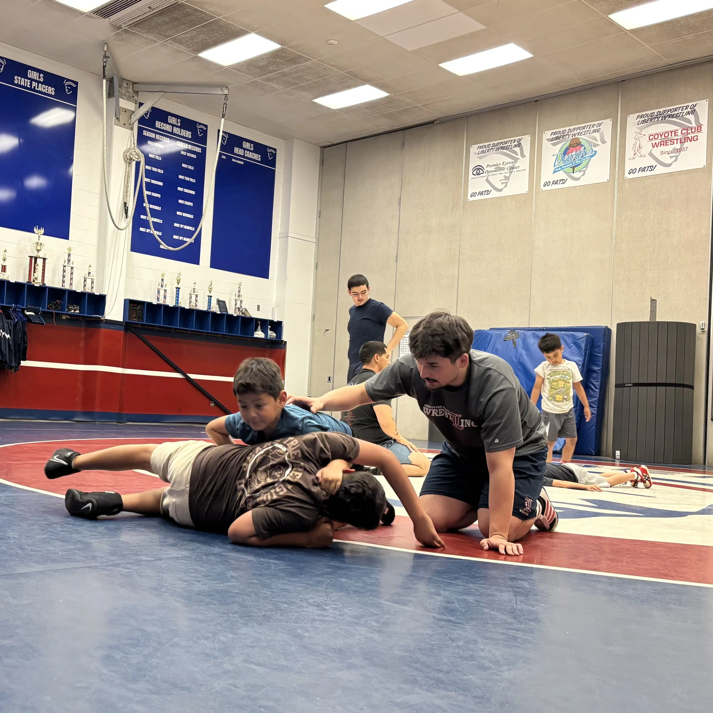 Young boys and a coach participating in a wrestling practice or class inside a gymnasium. The coach is demonstrating a move on a boy lying on the mat, while other boys observe. The gym has blue and red wrestling mats, trophies on shelves, and banners on the wall supporting Liberty Wrestling.