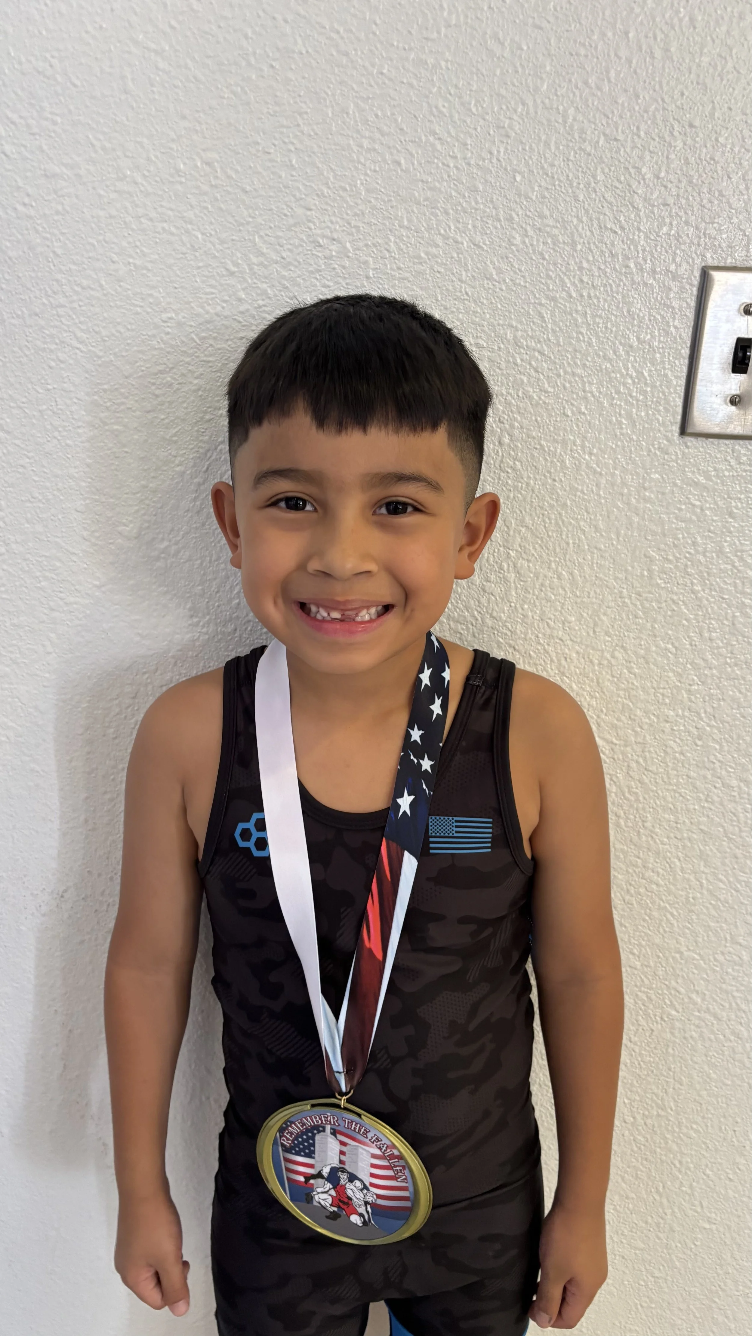 Young boy wearing a black camouflage athletic outfit and medals, smiling at the camera, standing against a plain white wall.