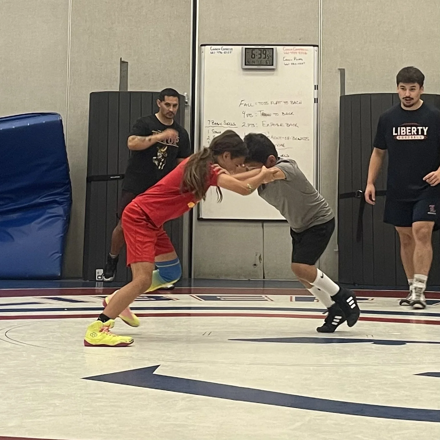 Two young men are wrestling on a gym floor while two others watch, with a whiteboard and digital clock in the background.