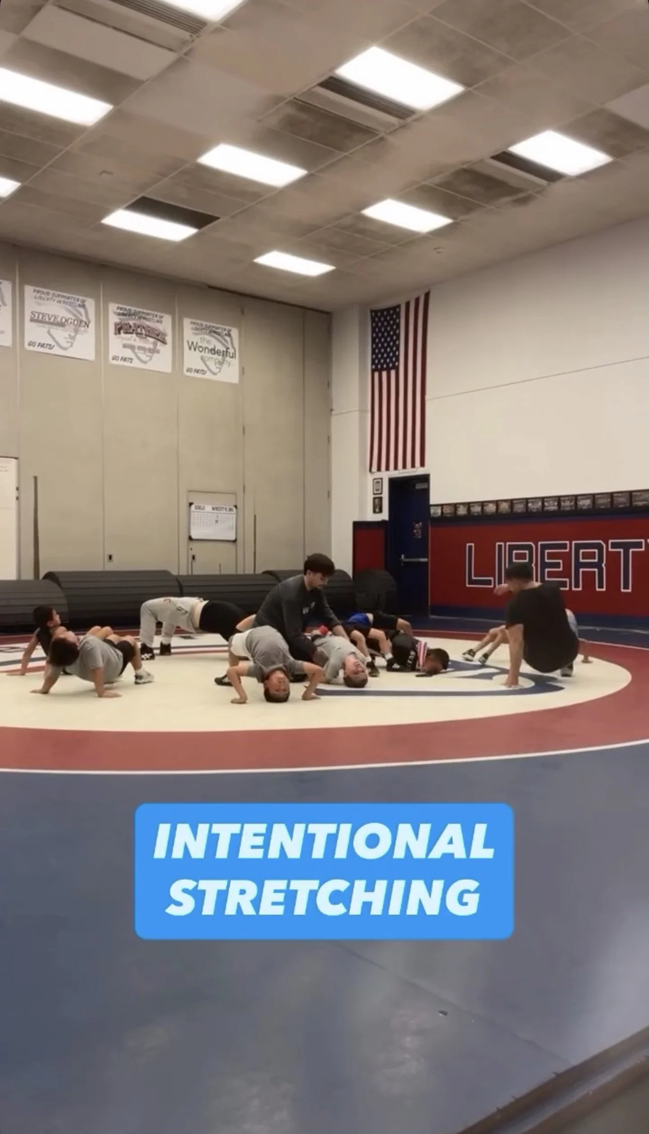 A group of children and adults practicing stretching exercises on a wrestling mat in a gymnasium. The gym has an American flag hanging on the wall, and signs promoting wrestling. A blue sign on the mat reads 'Intentional Stretching'.