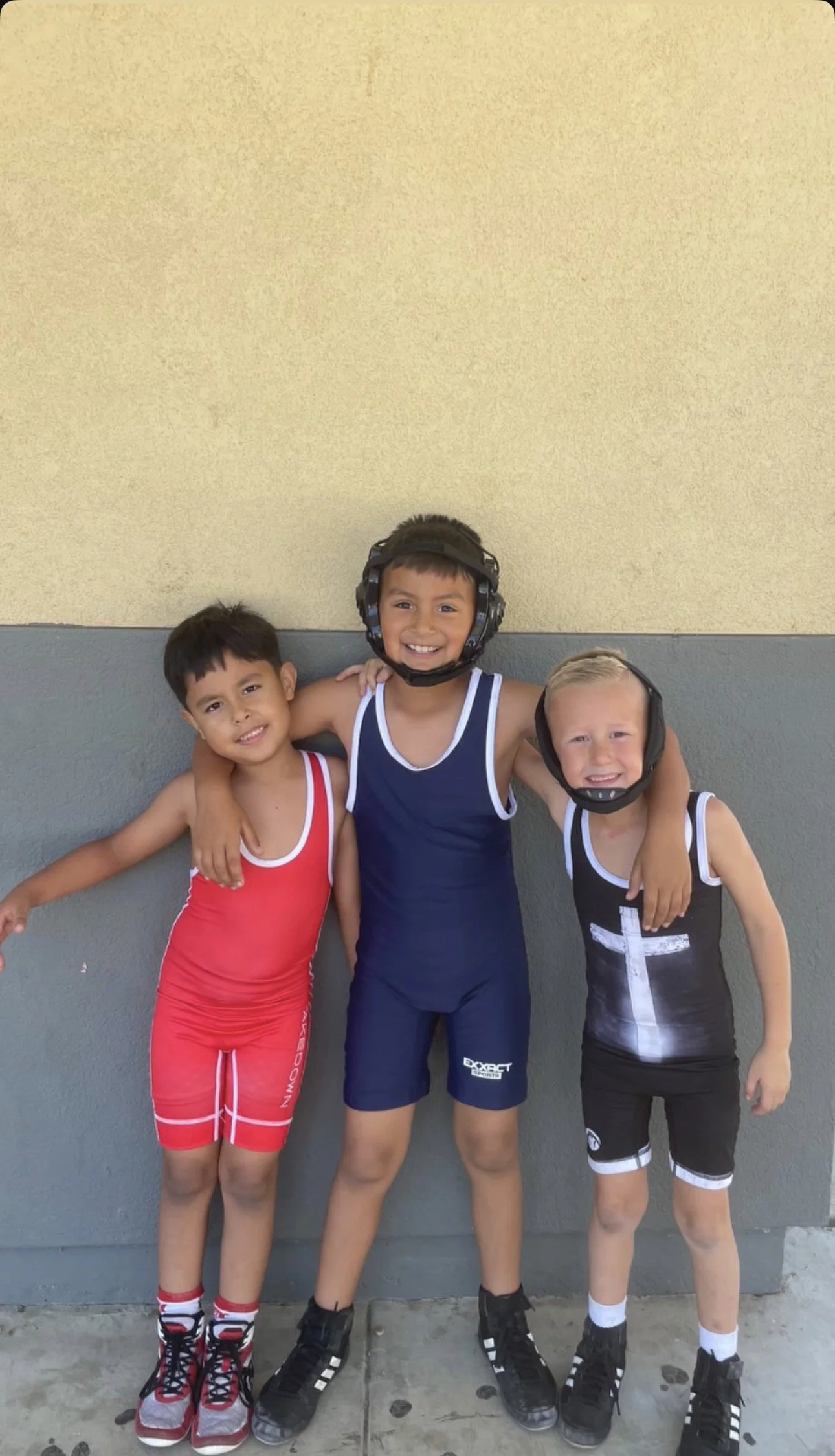 Three young boys in sports outfits, standing with arms around each other outside against a two-tone wall, smiling for the camera. The middle boy wears a helmet.