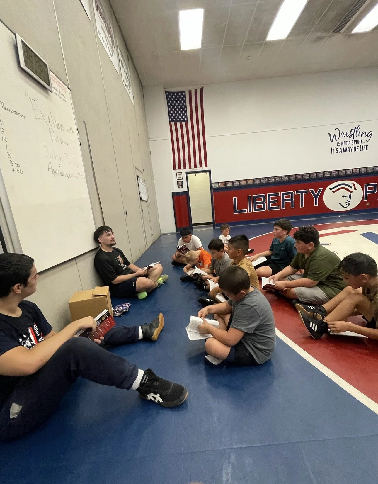 A group of young boys sitting on the floor in a wrestling gym, listening to an adult who is also sitting on the floor. The gym has an American flag on the wall, a Liberty logo, and a whiteboard with notes.