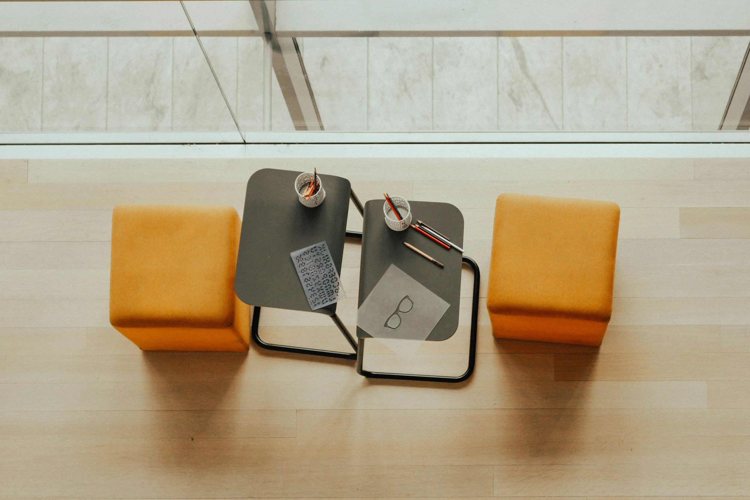 Top-down view of a waiting area with two yellow chairs, two small black tables, and various office supplies, including pencils, a paper with eyeglasses illustration, a window with a white frame, and a marble floor.
