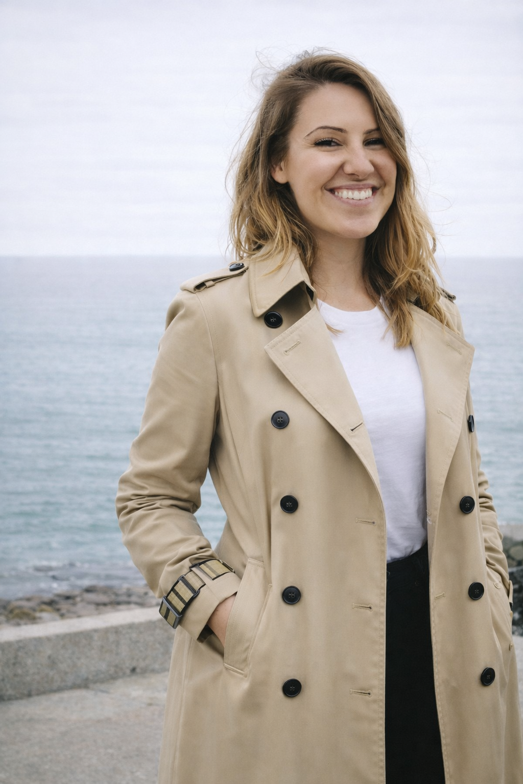 A smiling woman wearing a beige trench coat standing outdoors near the water with a cloudy sky in the background.