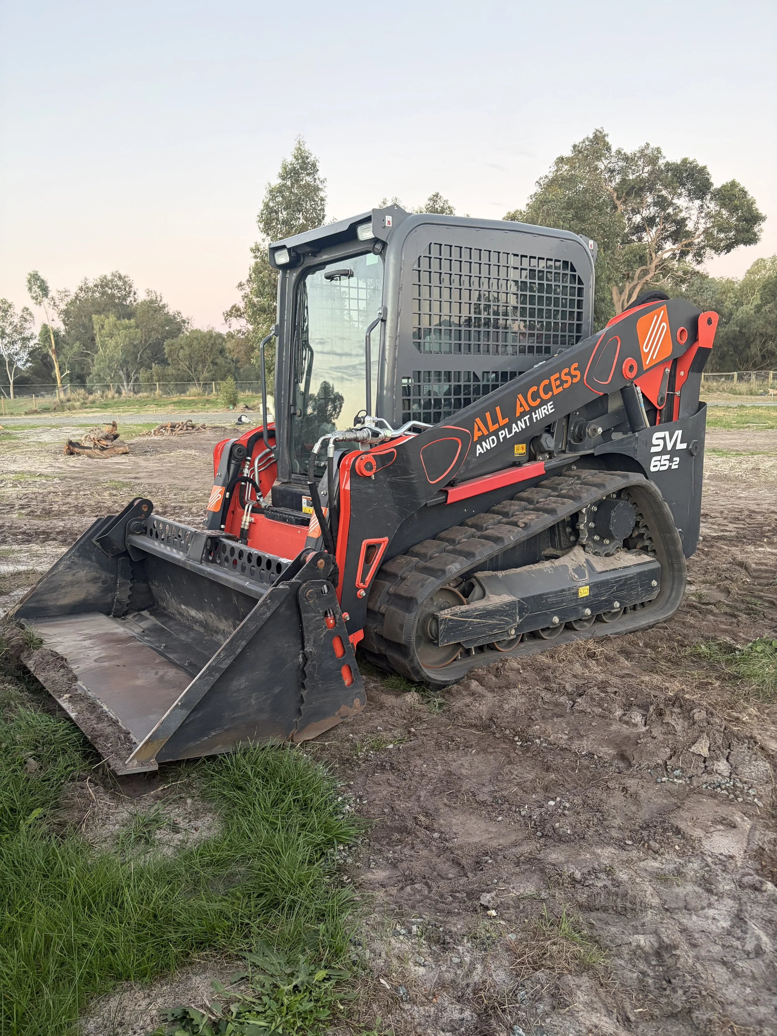 A small tracked excavator with a black and red exterior sitting on dirt and grass in an open outdoor area, with trees and a fence in the background.