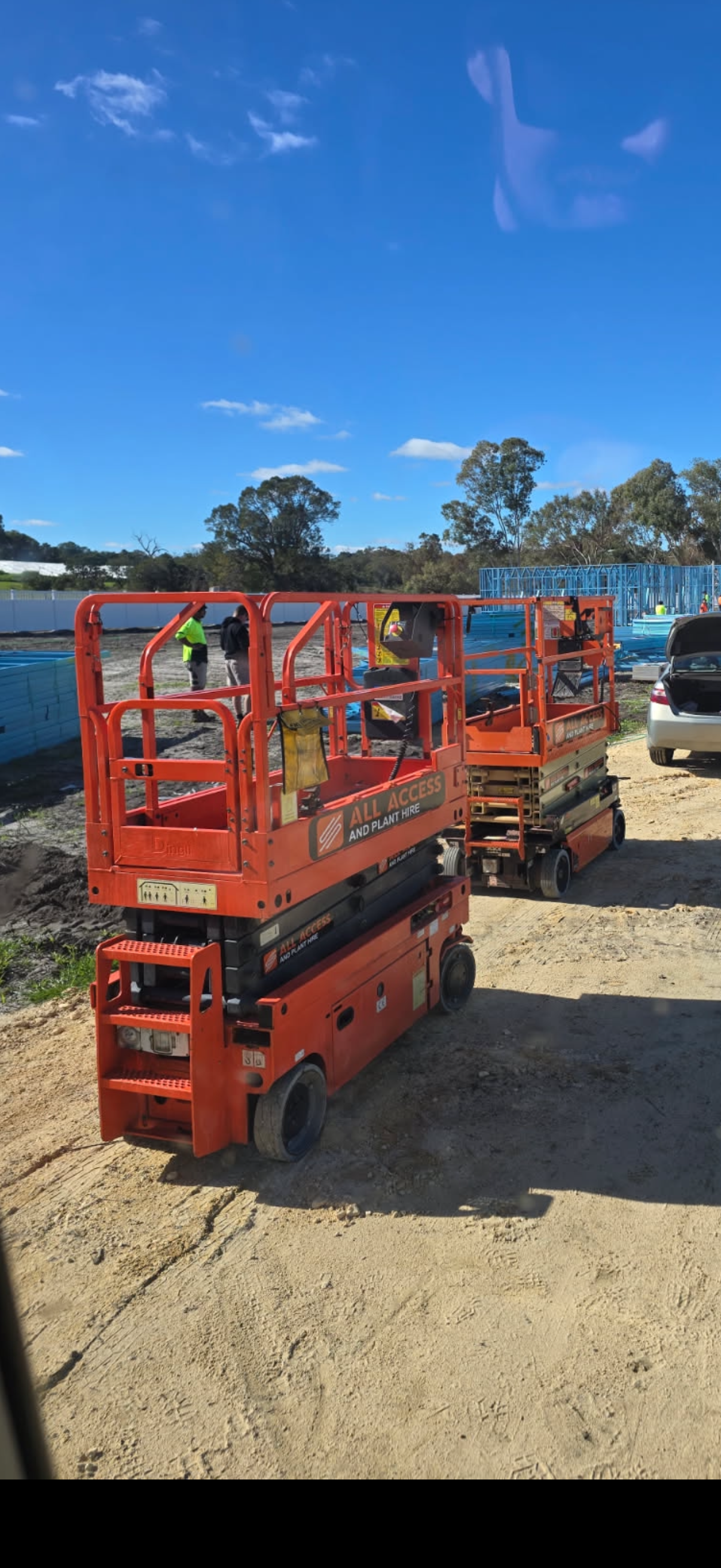 Construction site with orange scissor lifts, workers, blue framing for a building, and a car under a clear blue sky.