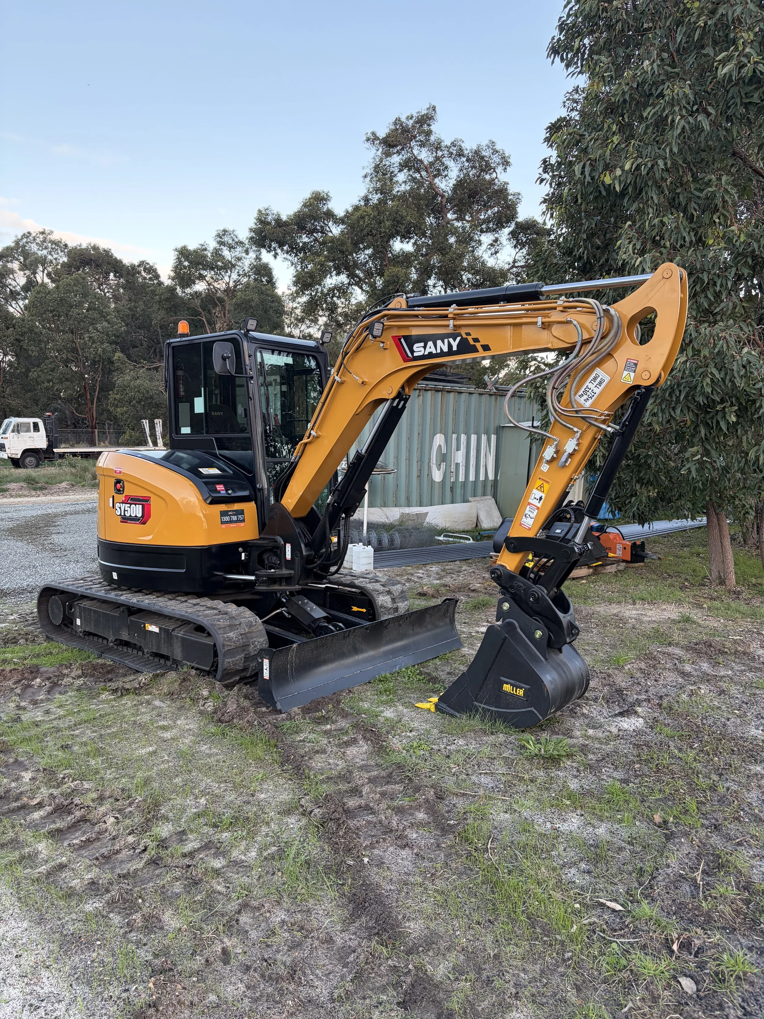 Yellow and black mini excavator with tracks on dirt ground, trees in background, blue sky.