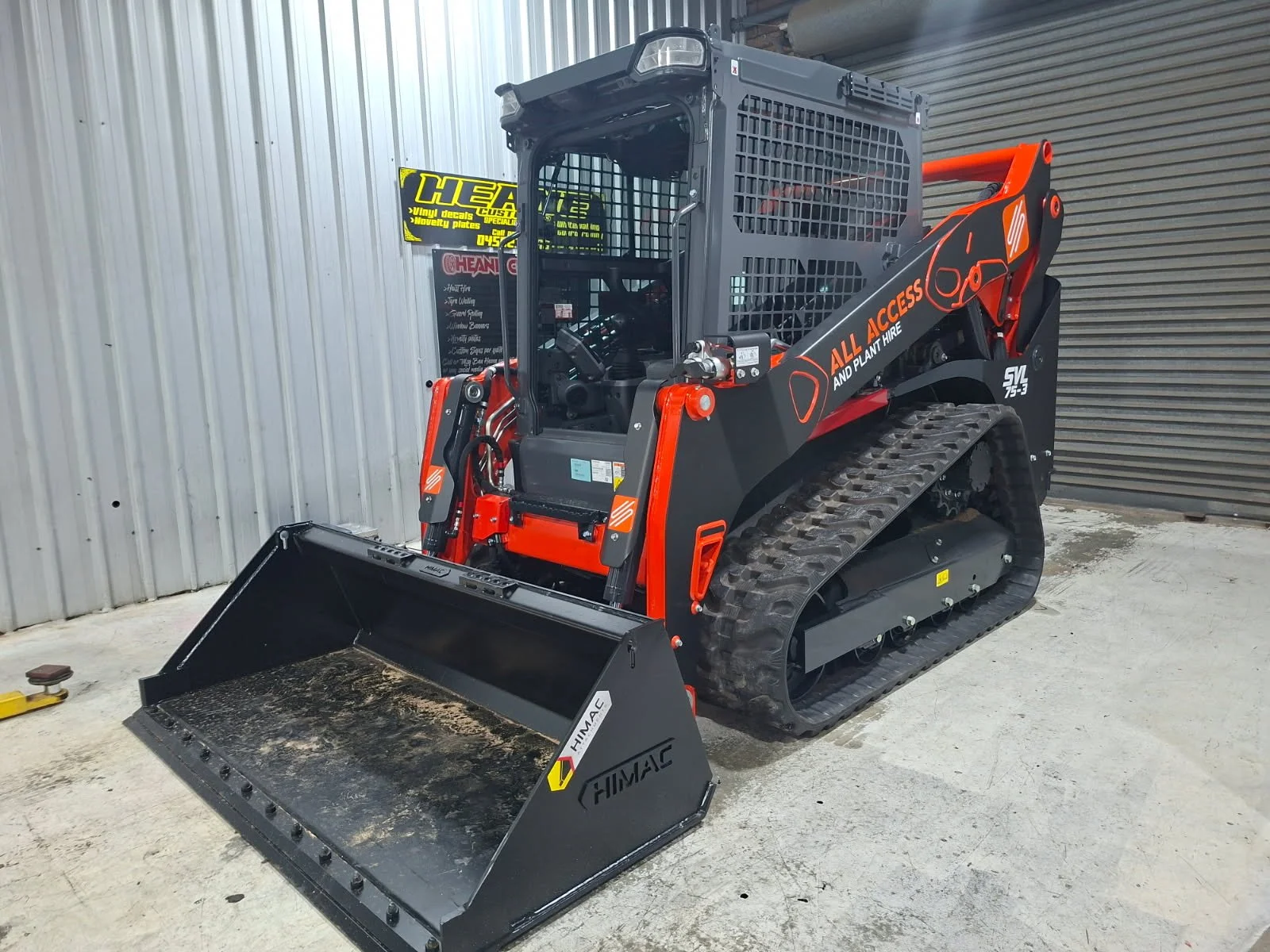 A tracked skid steer loader inside a warehouse with metal walls. The black and orange machine has a front bucket with dirt and debris, and branding that reads 'ALL ACCESS AND PLANT HIRE' and 'SVL 75-3'.