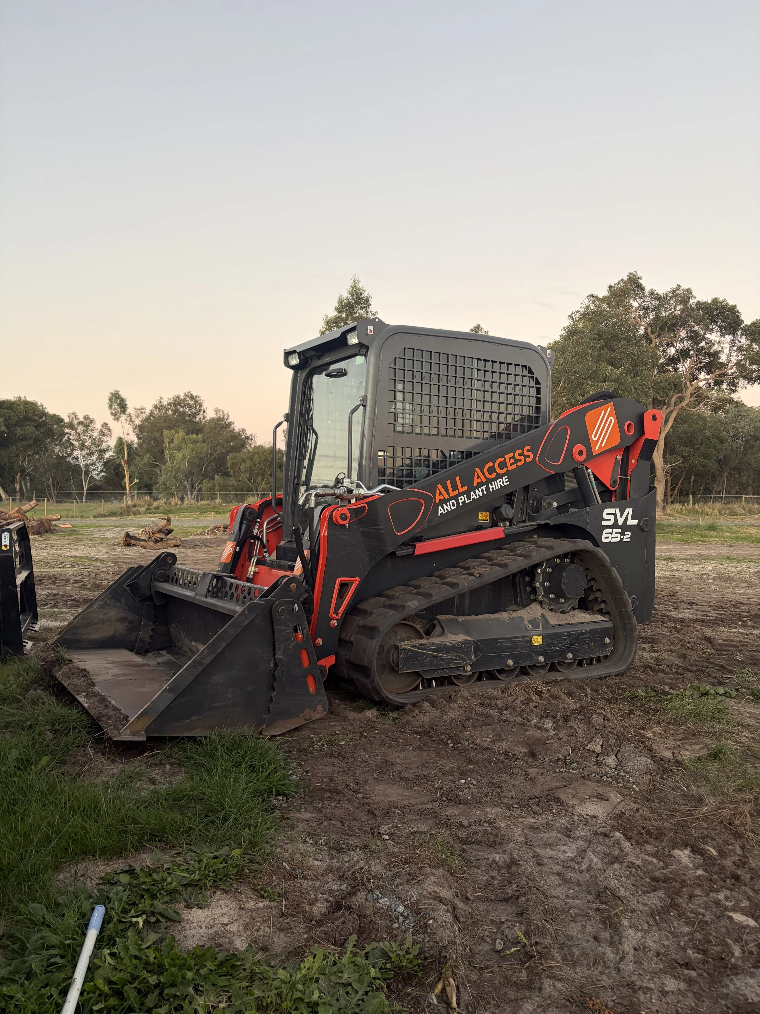 A compact excavator with the text 'ALL ACCESS AND PLANT HIRE' on its arm, parked on a dirt field with trees in the background during sunset.