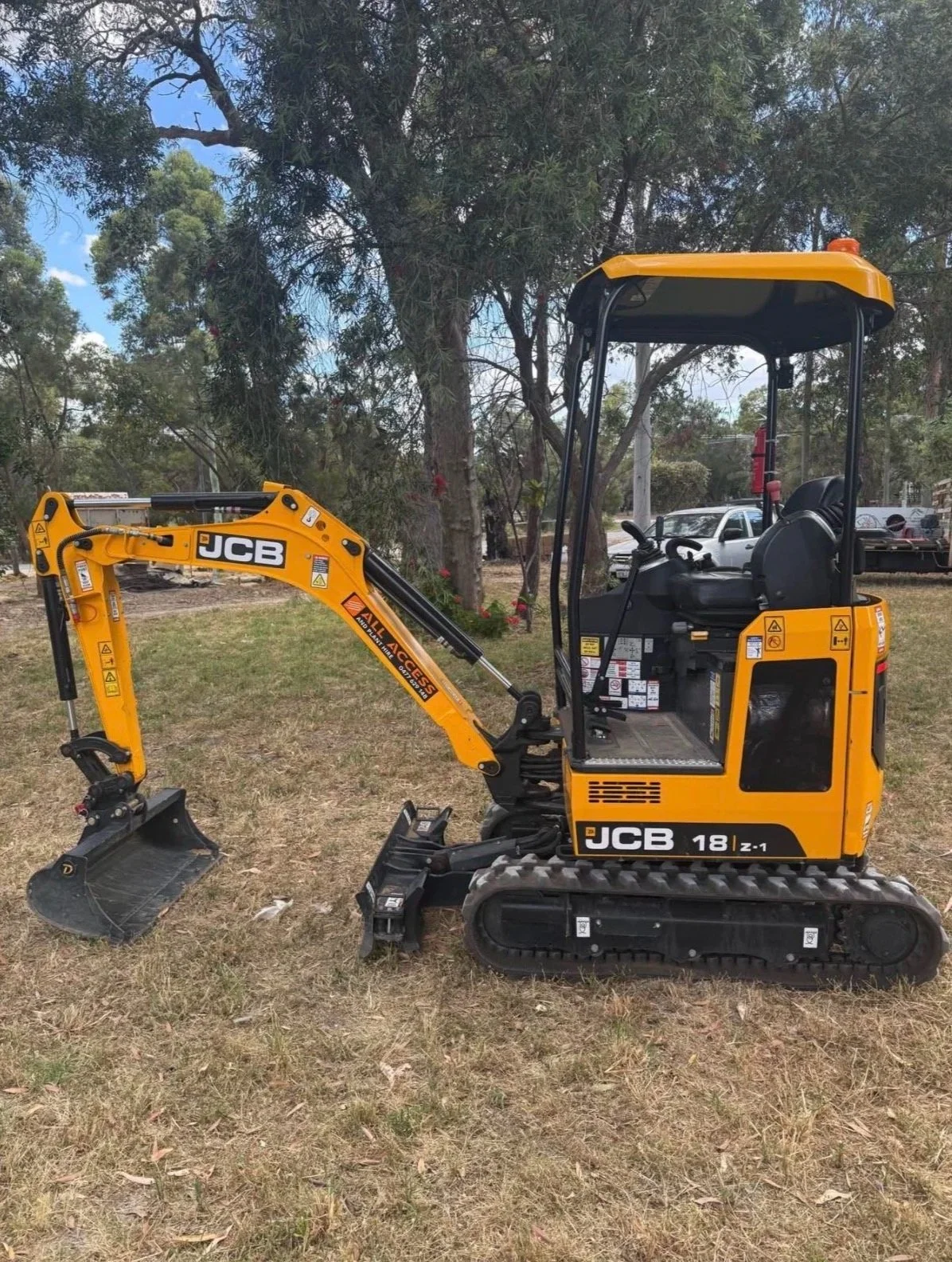 A yellow JCB mini excavator parked on a grass field with trees and cars in the background.