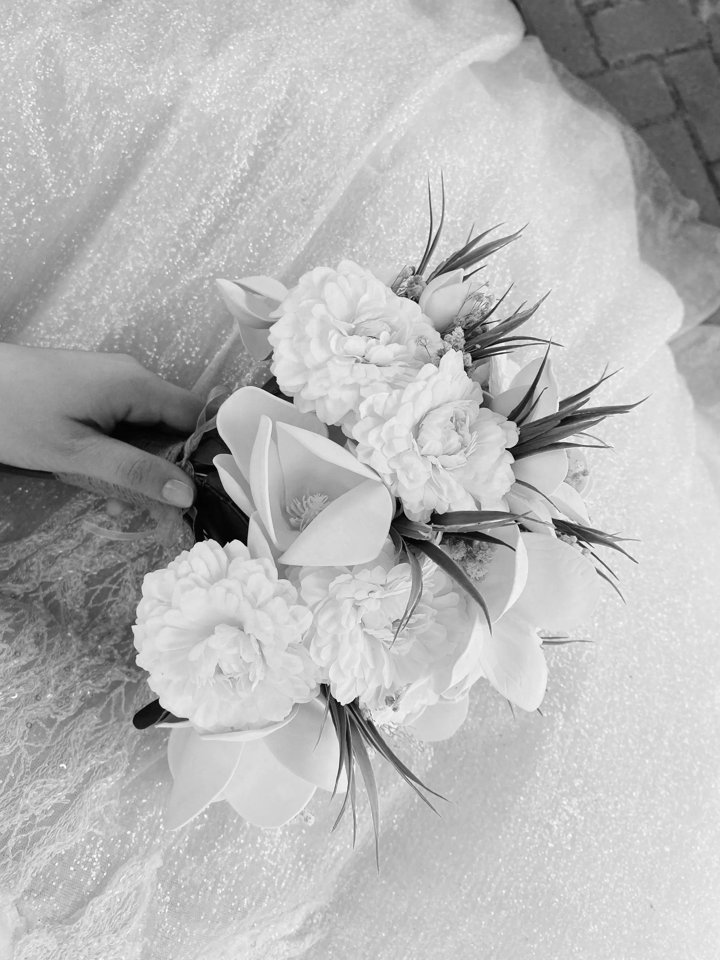 Black and white photo of a hand holding a flower bouquet with various flowers, including roses and lilies, against a glittery background.