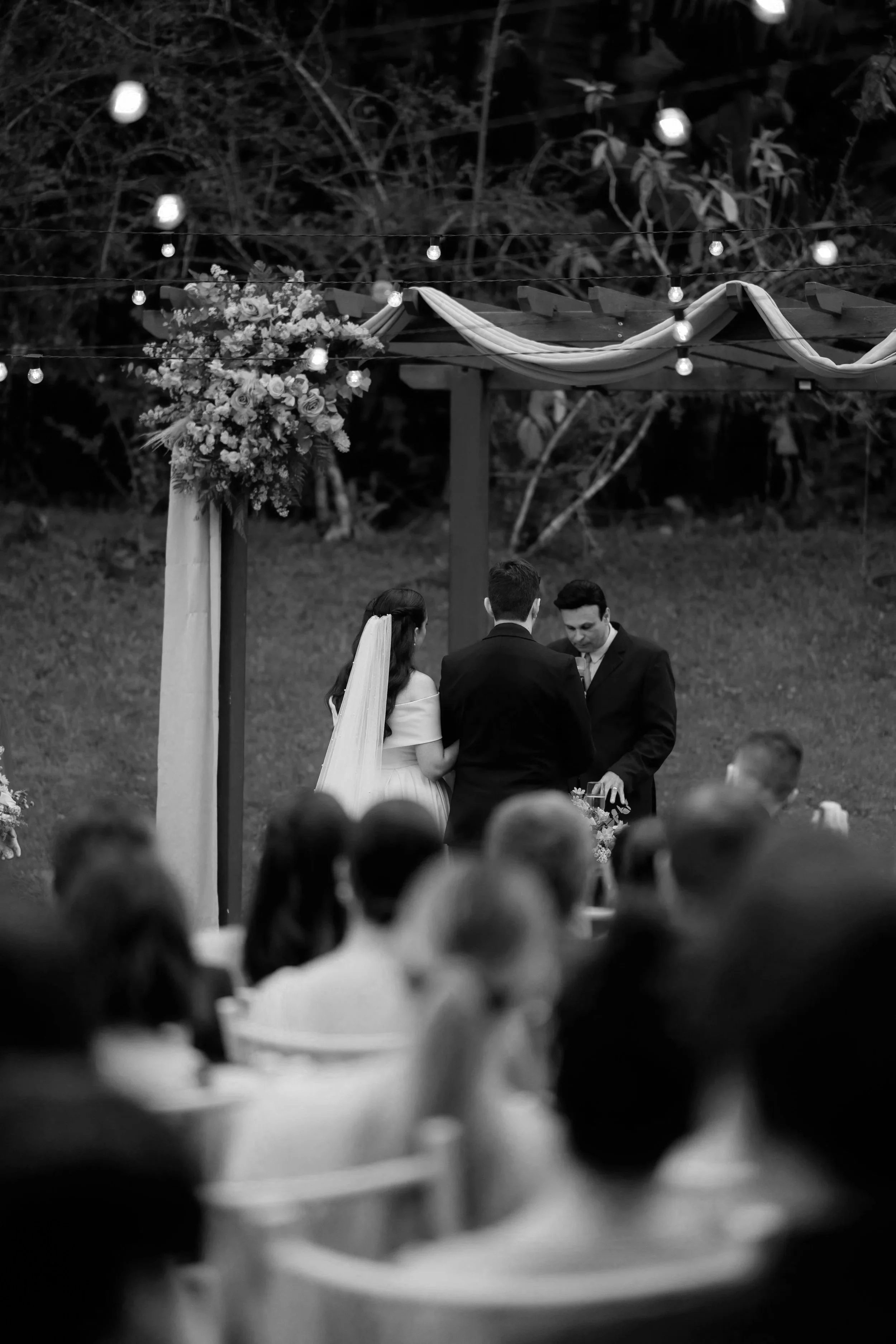 Black and white photo of a wedding ceremony outdoors. A couple stands before an officiant, with guests seated and watching. Decorations include string lights and a floral arrangement on a wooden arch. Trees and foliage are in the background.