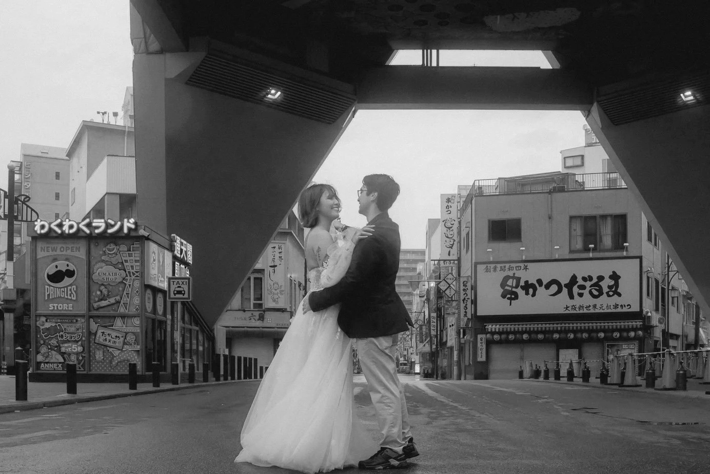 A couple in wedding attire dancing in the middle of a city street under a large elevated structure, with commercial signs and buildings in the background.