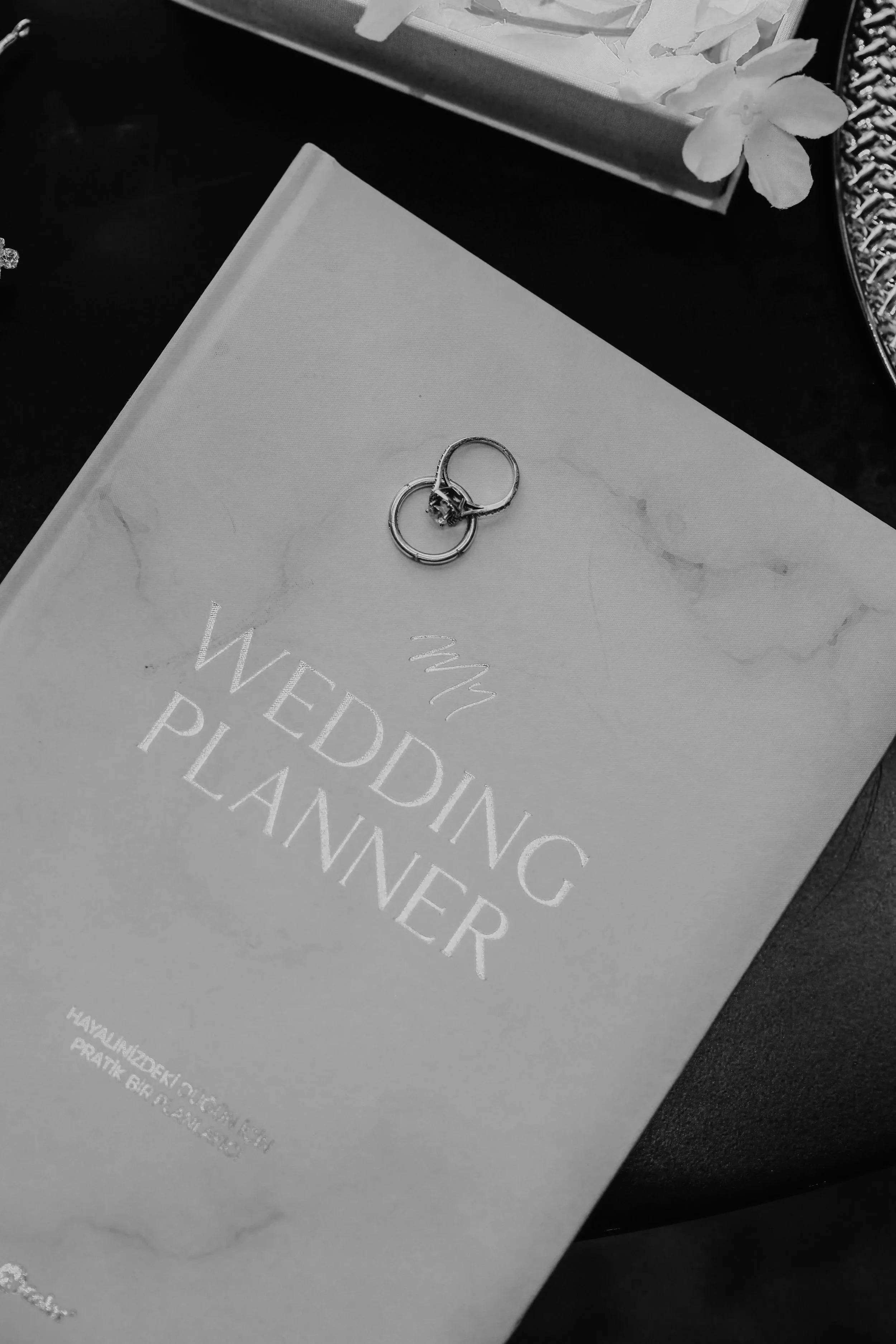 Two wedding rings, one with a large center stone, resting on a white book with partially visible text. In the background, there are white flowers in a flat rectangular container and a decorative silver tray with a chain design.