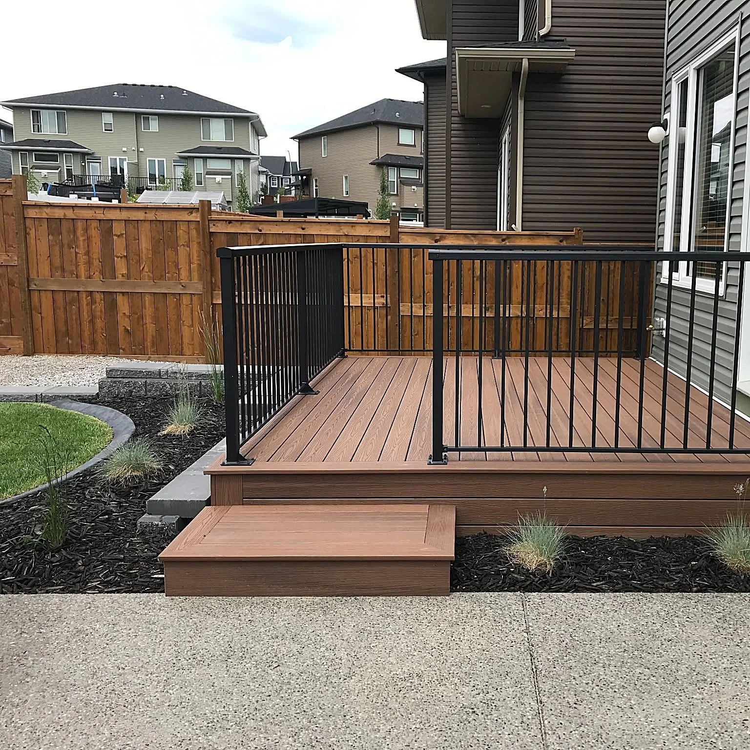 View of a backyard deck with a black metal railing, wooden steps, and a surrounding garden with plants and mulch. Several multi-story houses are visible in the background.