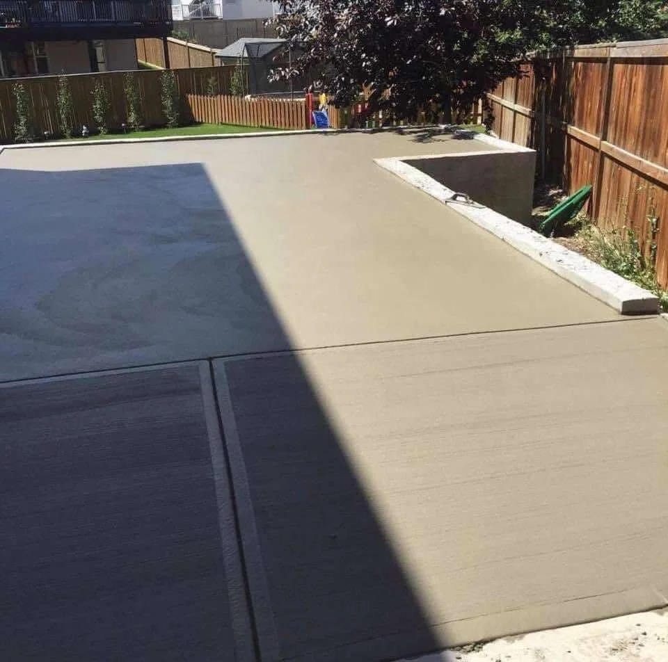 A backyard patio with a concrete surface, fenced with wooden panels, and a lawn area with a tree and backyard toys in the background.