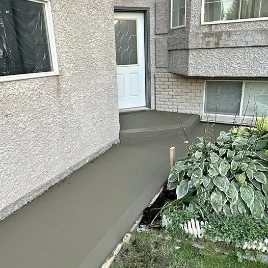Concrete sidewalk leading to a white front door with a decorative glass panel, surrounded by white-washed outdoor plants and a small garden bed.
