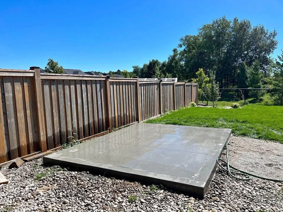 Freshly washed concrete patio outside a fenced backyard with grass and trees under a clear blue sky.