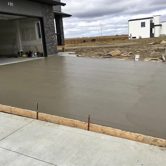 Concrete patio under construction in front of a modern house with an open garage. Construction materials and muddy ground visible in the background.
