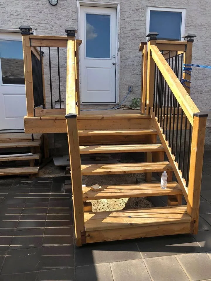 A wooden outdoor staircase with black metal railing leads up to a porch with a white door, attached to a stucco house. There is a water bottle on one of the steps and a piece of wood on another.
