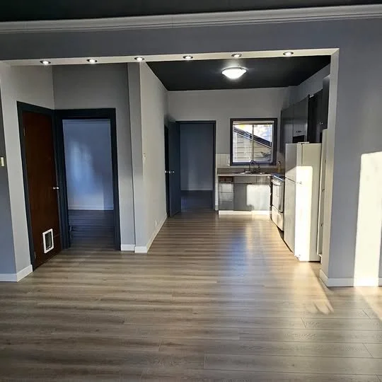 Empty living room and kitchen area with wood flooring, grey and white walls, a window, and appliances including a refrigerator and stove.