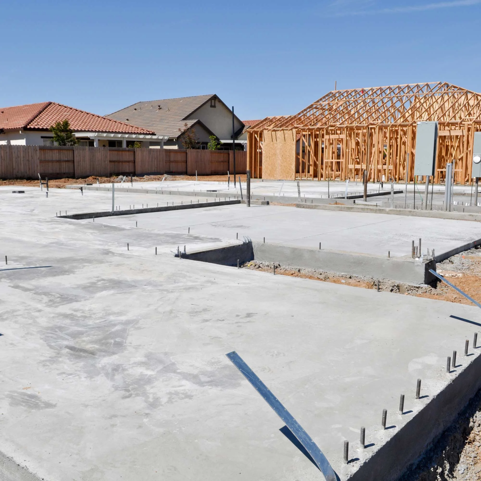 Construction site with unfinished wooden framing for a new building, with pre-existing houses in the background and a clear blue sky.