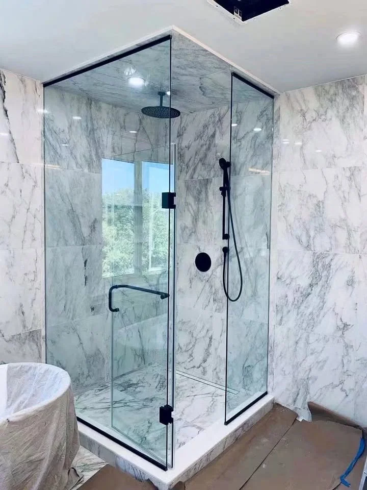A modern glass walk-in shower with marble walls and floor, black fixtures, and a ceiling-mounted rainfall showerhead in a bathroom with a window.