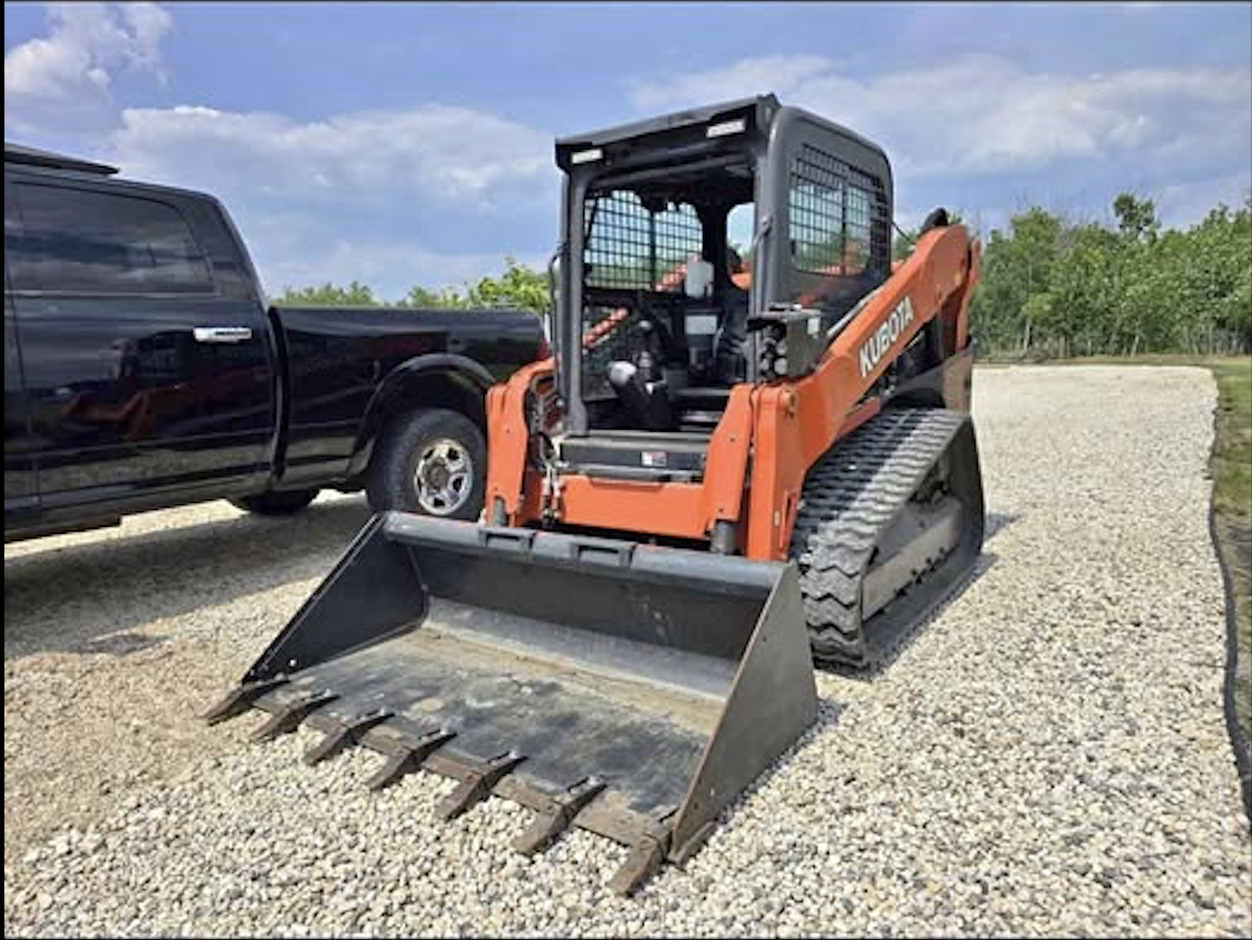 Orange Kubota compact track loader with a front bucket attachment parked on gravel, with a black pickup truck on the side and green trees under a partly cloudy blue sky.