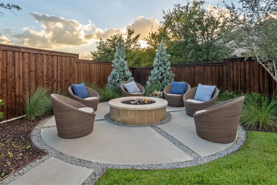 Outdoor backyard with a circular stone fire pit surrounded by six wicker chairs with cushions, enclosed by a wooden privacy fence, with trees and a sky with clouds in the background.
