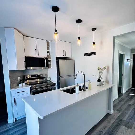 Modern kitchen with white cabinets, stainless steel refrigerator, microwave, and stove, featuring a white island with a sink, pendant lighting, and a gray wood floor.