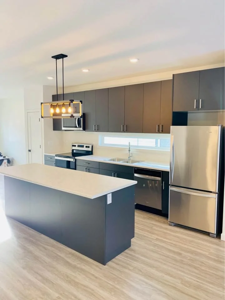 Modern kitchen with gray cabinets, stainless steel refrigerator, dishwasher, and oven, a large island with a light-colored countertop, and a decorative ceiling light fixture.