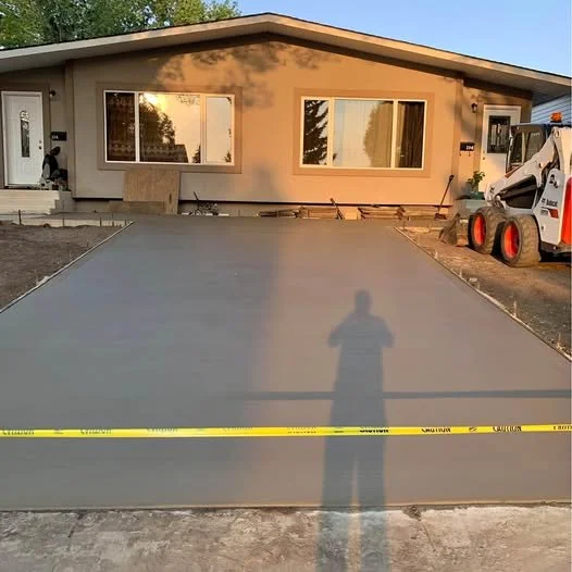 A freshly poured concrete driveway in front of a house during sunset. A person's shadow is visible on the driveway, and construction equipment is on the side.