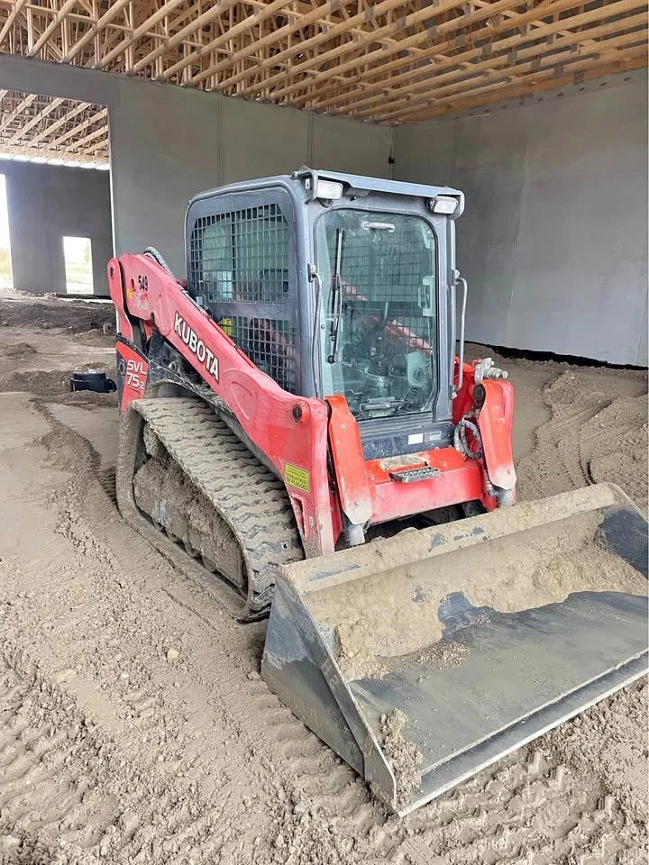 A Kubota compact track loader with a scoop bucket on a construction site, inside a building under construction with a wooden roof structure.