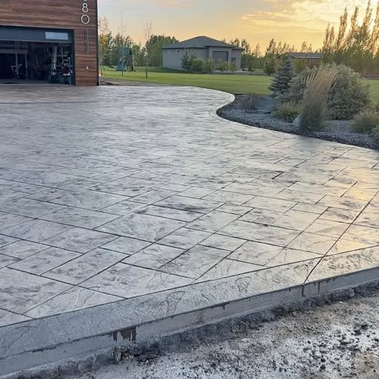 Concrete driveway with decorative stamped pattern, leading up to a garage door, with landscaping and trees in the background at sunset.