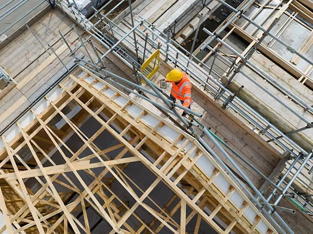 Construction worker wearing an orange safety vest and yellow helmet working on a wooden scaffolding structure at a construction site.